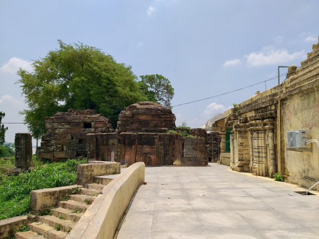 Ruinen eines Tempels mit alter Architektur, Wänden, Stufen, Geländern, Pflanzen, Bäumen und Drähten entlang einer Schotterstraße unter einem bewölkten Himmel.